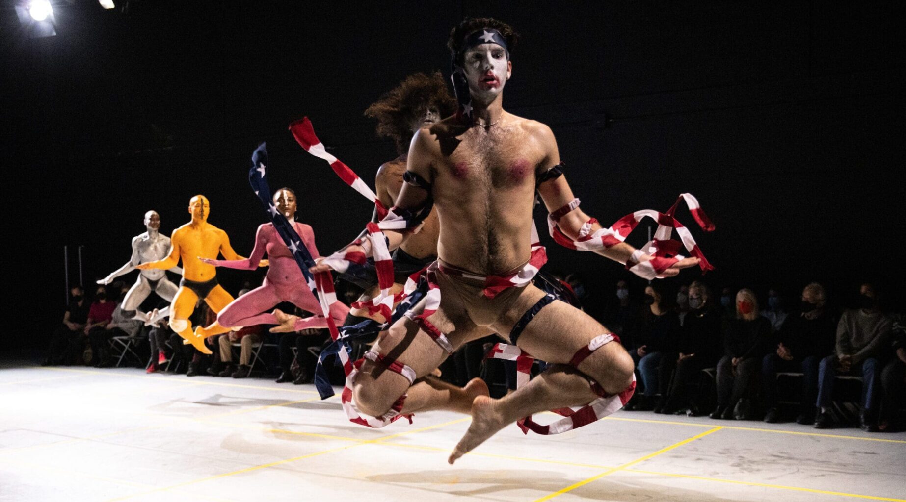 A line of dancers performs on a floor level white stage with the audience in the background. The performers are wearing body paint, minimal clothing, with the closest dancer wearing American flag ribbons on their body. The dancers are in a dynamic pose, caught jumping in the air with their legs tucked underneath themselves.