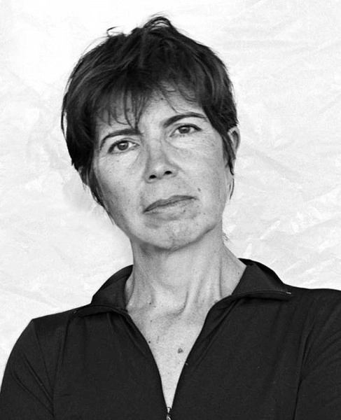 Black and white photo portrait of Elizabeth Diller. They are standing against a white stucco wall wearing a black collared shirt.