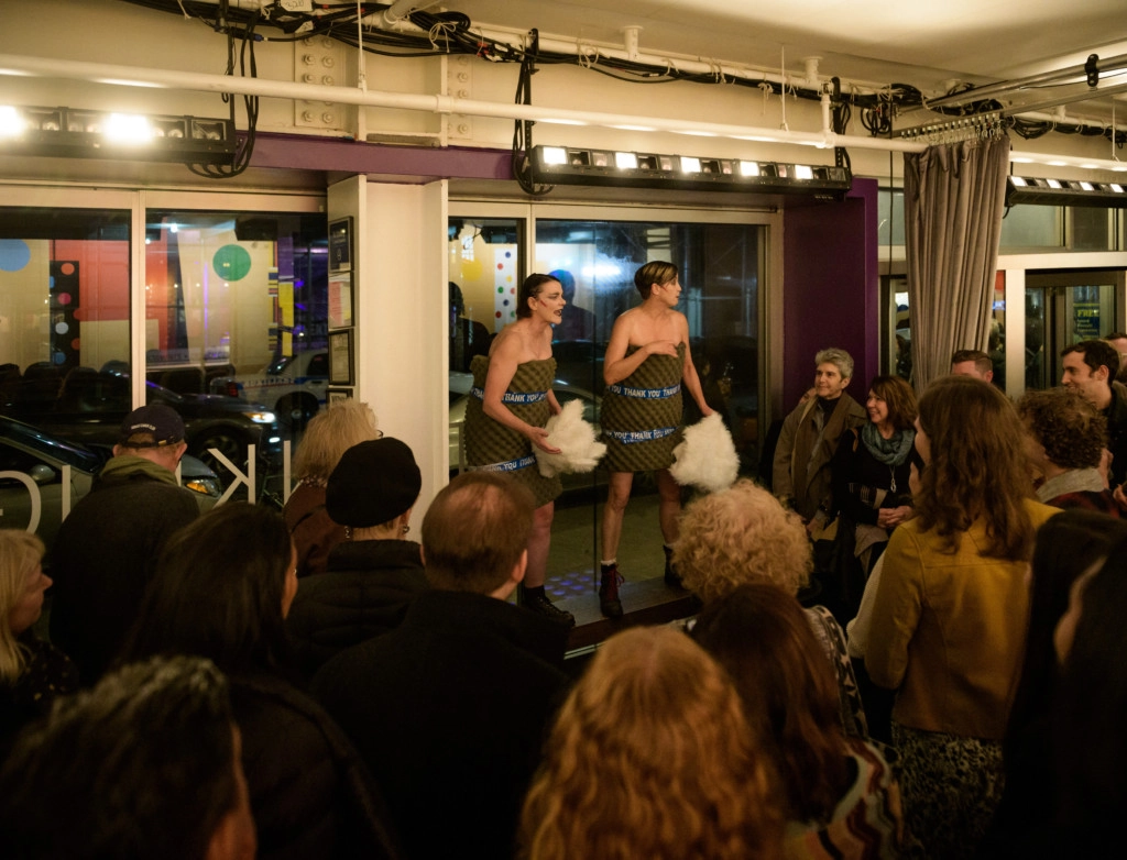 Two figures performing in a crowded room. They are both wearing eccentric eye makeup, their bodies wrapped in foam padding, and holding fluffy white objects. They are standing on a small bench facing the audience who looks on with their backs turned to the camera.