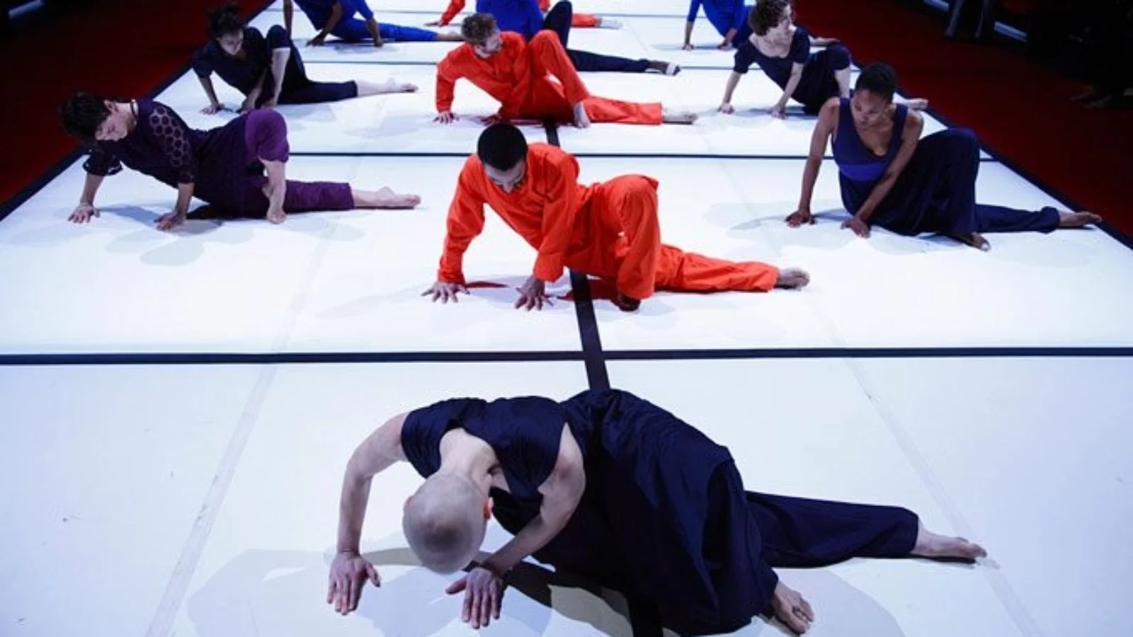 Three rows of dancers sit hunched over on a luminescent white stage