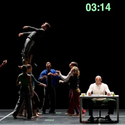 Bill T Jones sits at a desk on stage while dancers perform acrobatic positions behind him